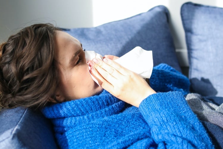 Woman in a blue sweater laying on the couch blowing her nose